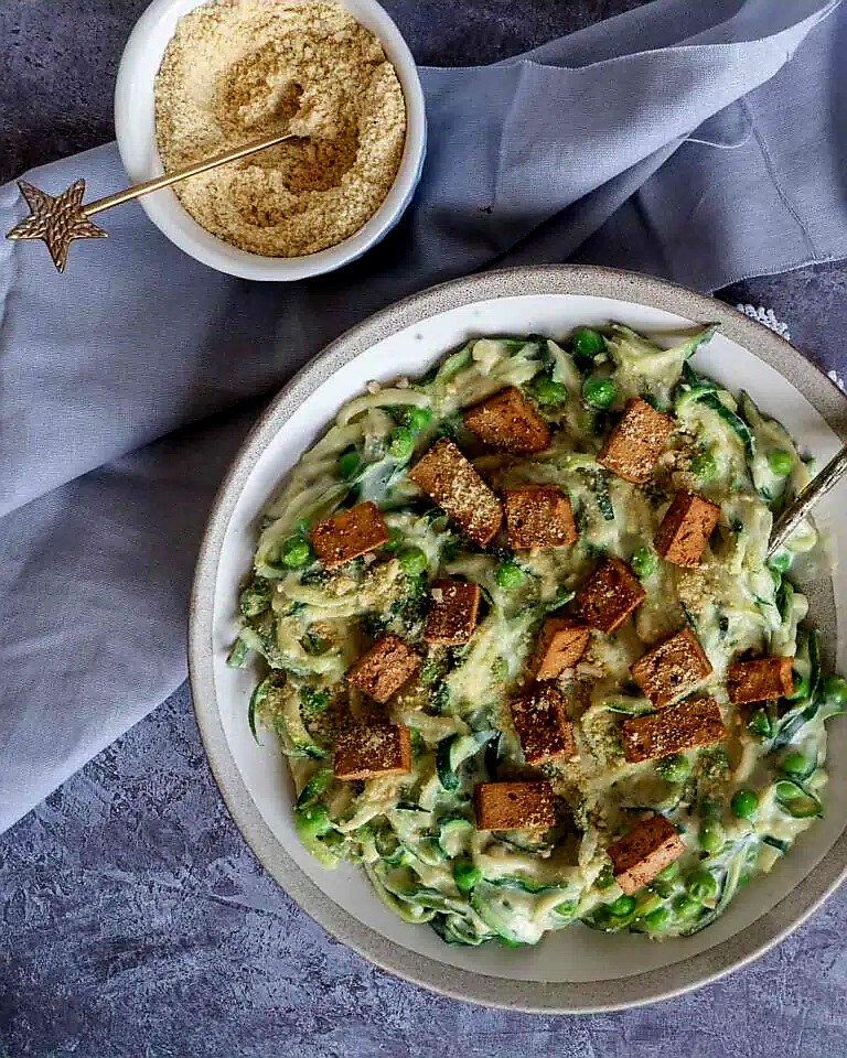 A bowl of creamy vegan carbonara pasta topped with crispy tofu bacon and peas, served with a side of vegan parmesan.